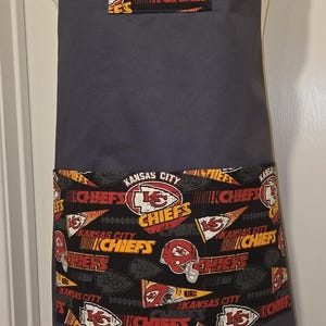 May include: A dark gray apron with a pocket and trim featuring the Kansas City Chiefs logo and text. The apron has a solid dark gray body and adjustable straps.