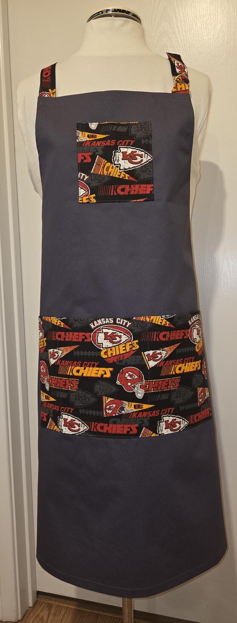 May include: A dark gray apron with a pocket and trim featuring the Kansas City Chiefs logo and text. The apron has a solid dark gray body and adjustable straps.
