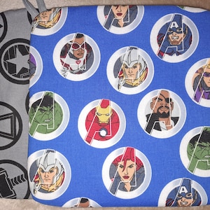 Puede incluir: Un agarrador de olla de tela azul con un patrón repetido de personajes de los Vengadores en círculos blancos. Los personajes incluyen Iron Man, Capitán América, Thor, Hulk, Viuda Negra, Ojo de Halcón, y más. El agarrador de olla está rodeado de una tela gris con un patrón repetido de logotipos de los Vengadores.