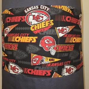May include: A dark gray apron with a black pocket featuring a repeating pattern of Kansas City Chiefs logos, helmets, and text in red, yellow, and white. The apron is made of a durable fabric.