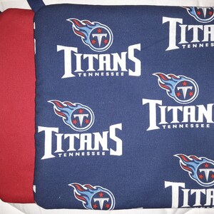Pode incluir: Um conjunto de dois pegadores de panela, um vermelho e um azul. O pegador azul tem um padrão repetido do logotipo do Tennessee Titans, que apresenta um "Titans" branco com "Tennessee" abaixo, uma chama azul e um "T" branco com três estrelas.