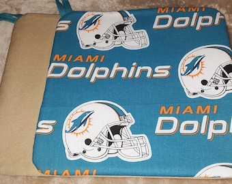 Puede incluir: Dos agarraderas cuadradas. Una es de color beige, la otra es de color turquesa con un patrón repetido de cascos de fútbol de los Miami Dolphins y el nombre del equipo en naranja y blanco. Cada agarradera tiene un lazo turquesa para colgar.