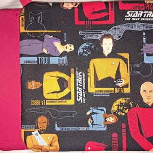 May include: A square pot holder with a dark grey background featuring Star Trek characters and logos. The pot holder has a bright pink border and hanging loop. The text "STAR TREK THE NEXT GENERATION" is visible.
