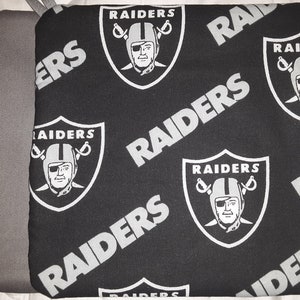 May include: Black and white fabric with a repeating pattern of the Las Vegas Raiders logo. The logo features a silver helmet with a black face mask and crossed swords. The words "RAIDERS" are repeated throughout the fabric.