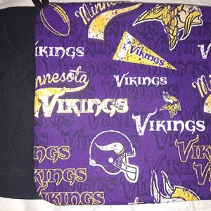 Puede incluir: Un agarrador morado y negro con el tema de los Minnesota Vikings. El lado morado tiene un patrón repetido del logotipo del equipo, el casco y la palabra "Vikings" en blanco y amarillo. El otro lado es negro liso.