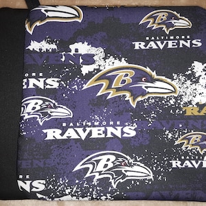 Puede incluir: Un agarrador de ollas cuadrado con el logotipo y el nombre de los Baltimore Ravens en blanco y dorado sobre un fondo morado y negro. Tiene un lazo negro para colgar.