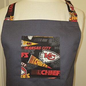 May include: A gray apron with a pocket featuring a Kansas City Chiefs design. The pocket has a black background with the team's logo and text in red, yellow, and white. The straps also have the team's logo.