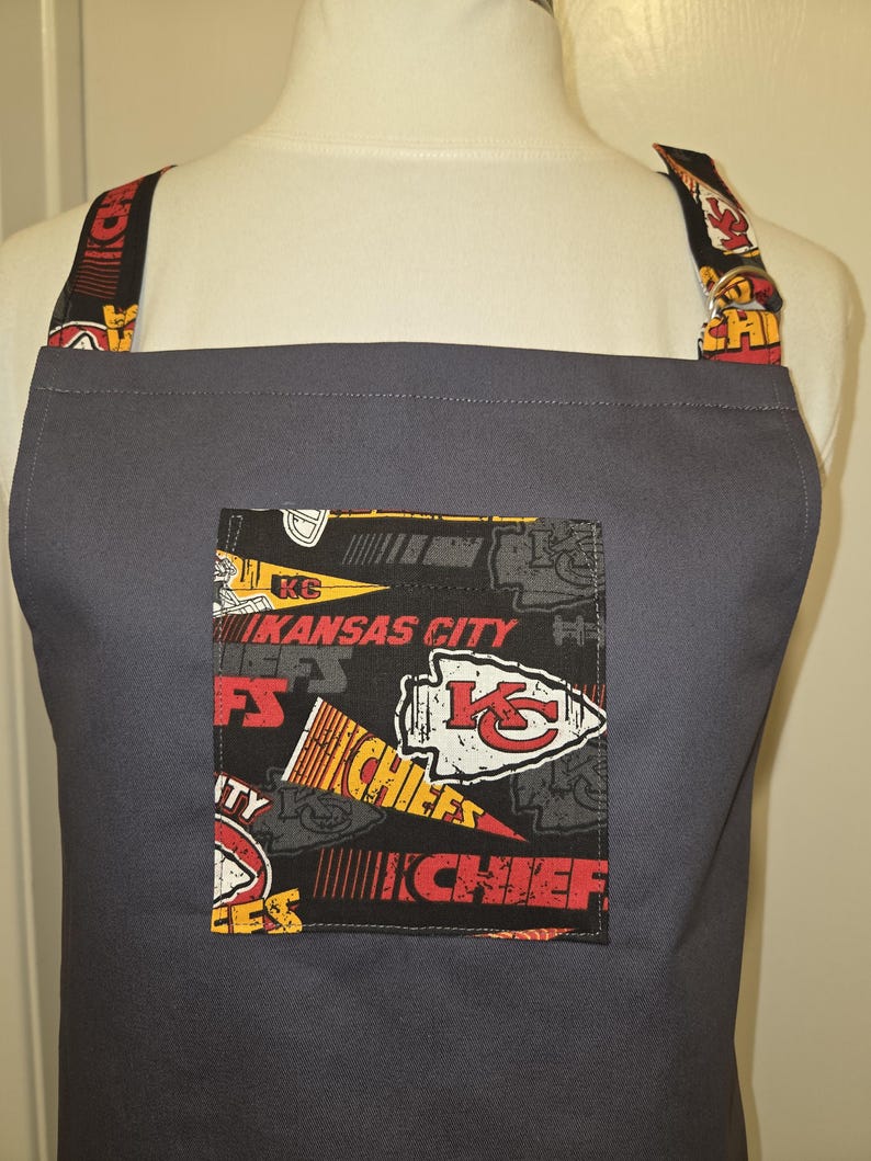 May include: A gray apron with a pocket featuring a Kansas City Chiefs design. The pocket has a black background with the team's logo and text in red, yellow, and white. The straps also have the team's logo.