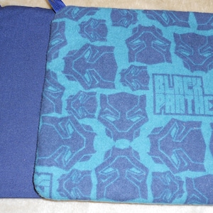 Puede incluir: Una bolsa de tela azul y turquesa con un diseño de Black Panther. La bolsa tiene una cremallera y una lengüeta de cierre negra. La bolsa está hecha de una tela suave y afelpada.