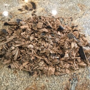 May include: A pile of brown wood chips with some black charred pieces. The chips are on a light brown and gray speckled surface.