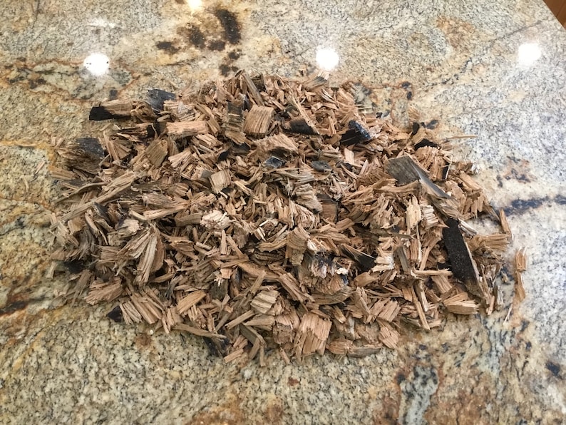 May include: A pile of brown wood chips with some black charred pieces. The chips are on a light brown and gray speckled surface.