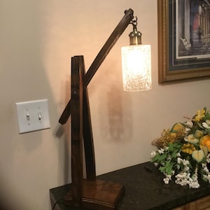 May include: A handcrafted wooden desk lamp with a textured glass shade. The lamp features a dark brown wooden frame and a brass-colored fixture. The shade is cylindrical and emits a warm, inviting light. The lamp is placed on a dark countertop.