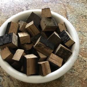May include: A white ceramic bowl filled with small, charred wood cubes. The cubes are various shades of brown and black, and some have a slightly charred appearance.