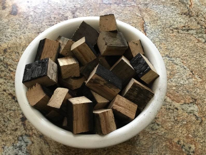 May include: A white ceramic bowl filled with small, charred wood cubes. The cubes are various shades of brown and black, and some have a slightly charred appearance.