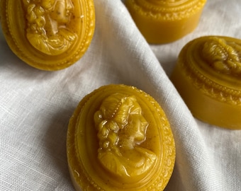 May include: Four yellow cameo soaps with a profile of a woman. The soaps are oval and round, with detailed relief carvings. The soaps are on a white, textured surface.