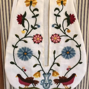 May include: White apron with embroidered floral and bird design. The apron features yellow, red, pink, and blue flowers, with two brown birds at the bottom. The design is symmetrical, with a vase in the centre.