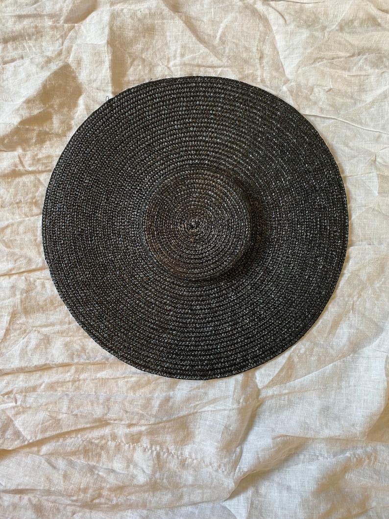 K&ouml;nnte beinhalten: Ein breitkrempiger, schwarzer Sonnenhut mit kreisf&ouml;rmigem Design. Der Hut besteht aus gewebtem Material und hat eine leicht erh&ouml;hte Krone. Der Hut soll vor der Sonne sch&uuml;tzen.