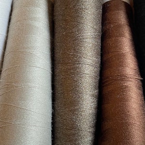 May include: Close-up of five spools of thread in varying colours. The colours include white, light grey, dark grey, brown, and black. The thread is tightly wound around the spools, creating a textured surface. Ideal for sewing and crafting projects.