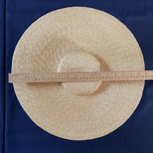 K&ouml;nnte beinhalten: Ein beigefarbener Sonnenhut mit breiter Krempe und kreisf&ouml;rmigem Muster. Der Hut hat einen Krempen-Durchmesser von ca. 43 cm. Der Hut besteht aus gewebtem Material und liegt auf einem dunkelblauen Hintergrund.