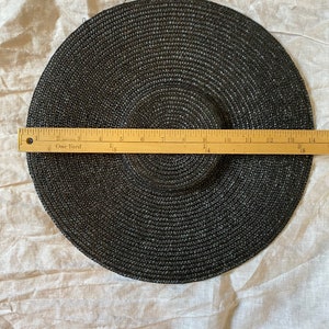 K&ouml;nnte beinhalten: Ein schwarzer Sonnenhut mit breiter Krempe, ca. 35,5 cm Durchmesser. Der Hut ist aus gewebtem Material gefertigt und hat ein kreisf&ouml;rmiges Design. Ein Holzlineal liegt zur Messung auf dem Hut.