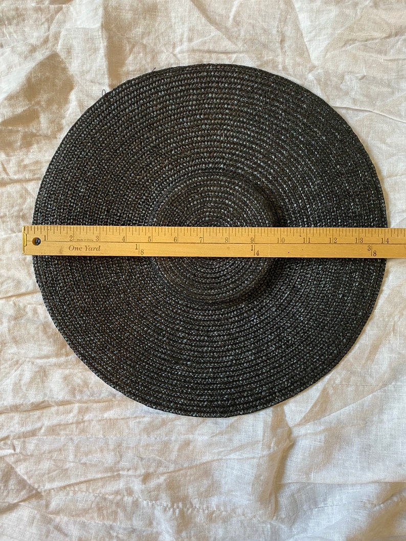 K&ouml;nnte beinhalten: Ein schwarzer Sonnenhut mit breiter Krempe, ca. 35,5 cm Durchmesser. Der Hut ist aus gewebtem Material gefertigt und hat ein kreisf&ouml;rmiges Design. Ein Holzlineal liegt zur Messung auf dem Hut.