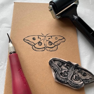 May include: A brown notebook with a black moth print, a red Speedball carving tool, a black brayer, and a carved moth stamp. The tools are arranged on a white surface, suggesting a printmaking or crafting setup.