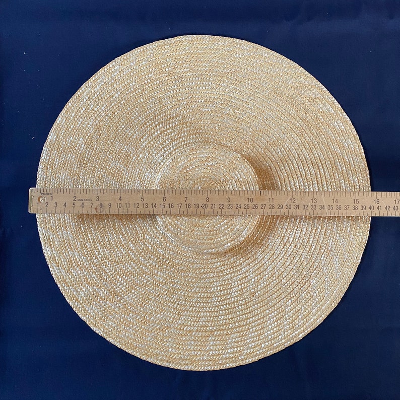 K&ouml;nnte beinhalten: Beigefarbener Strohhut mit breiter Krempe und kreisf&ouml;rmigem Design. Der Hut hat einen Krempen-Durchmesser von etwa 43 cm. Ein Holzlineal liegt zur Messung auf dem Hut.