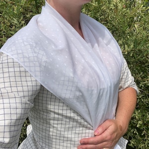 May include: A white scarf with a subtle dotted pattern draped over a checkered shirt. The shirt has a black and white grid pattern. The outfit is completed with a white bonnet.