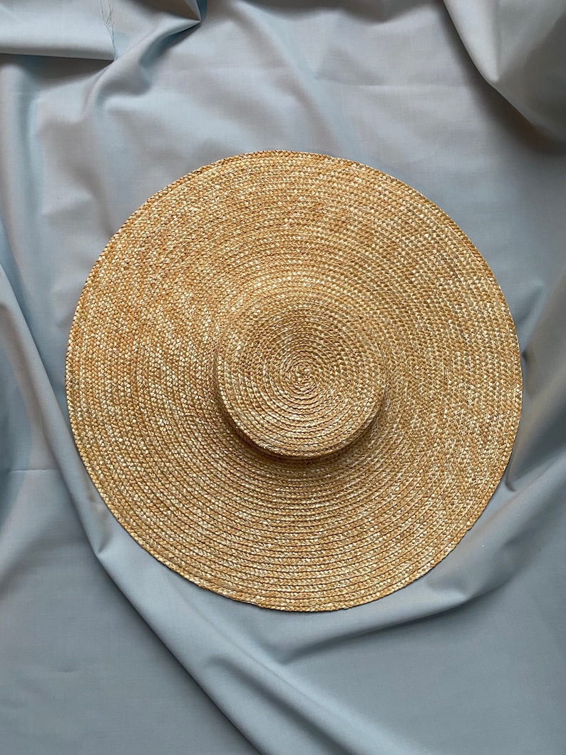K&ouml;nnte beinhalten: Ein breitkrempiger, nat&uuml;rlicher Strohhut mit kreisf&ouml;rmiger Krone und breiter, kreisf&ouml;rmiger Krempe. Der Hut ist aus gewebtem Stroh in Beige- und Beiget&ouml;nen gefertigt. Der Hut ist vor einem hellblauen Stoffhintergrund platziert.