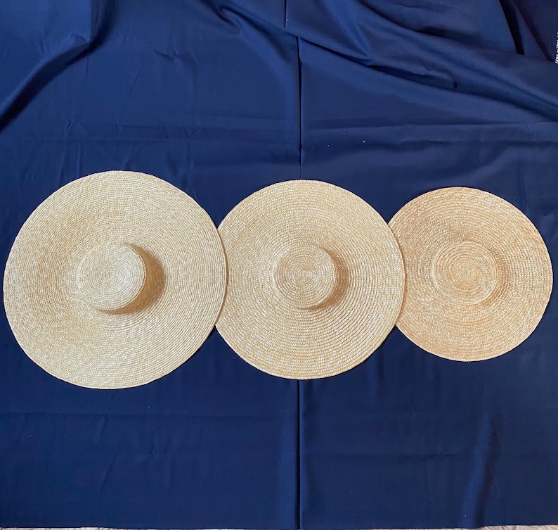 K&ouml;nnte beinhalten: Drei breitkrempige Strohh&uuml;te in Naturbeige sind auf einem dunkelblauen Hintergrund angeordnet. Die H&uuml;te haben eine kreisf&ouml;rmige Krone und eine breite, kreisf&ouml;rmige Krempe, ideal f&uuml;r den Sonnenschutz. Die H&uuml;te variieren leicht in der Gr&ouml;&szlig;e.