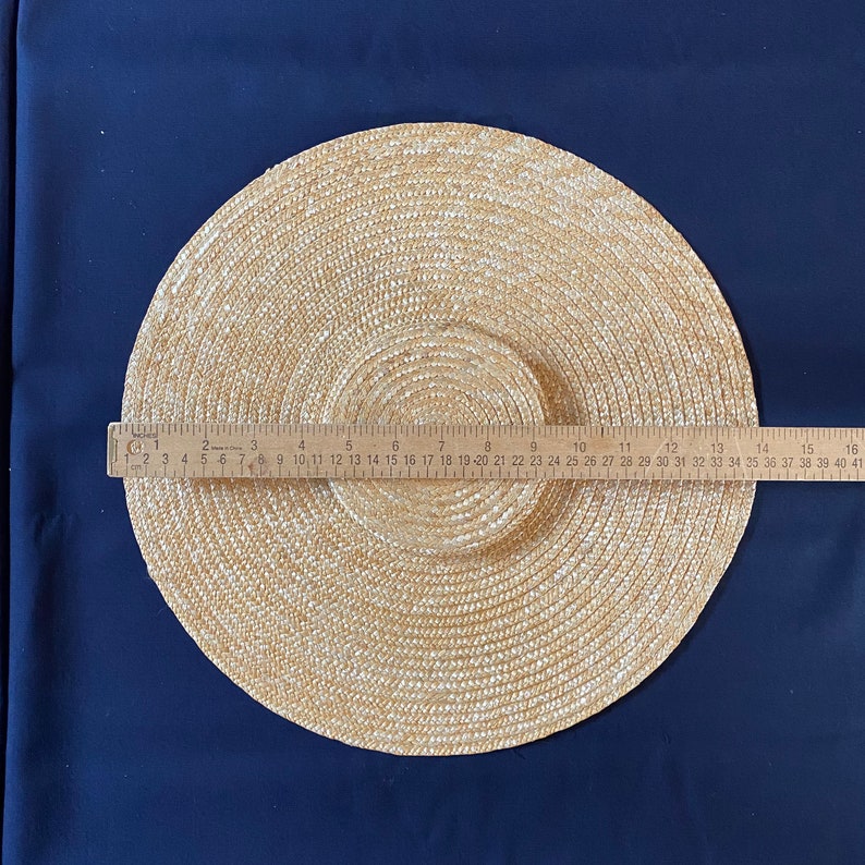 K&ouml;nnte beinhalten: Ein Sonnenhut aus Stroh mit breiter Krempe, etwa 40 cm im Durchmesser, wird vor einem dunkelblauen Hintergrund gezeigt. Der Hut besteht aus gewebtem Stroh und hat eine kreisf&ouml;rmige Krone und Krempe. Ein Lineal liegt zur Messung auf dem Hut.
