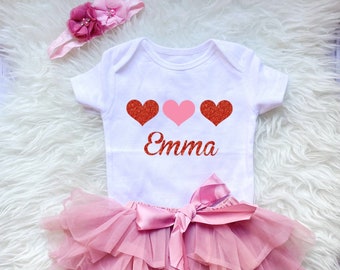 May include: A white baby onesie with three glitter hearts, one pink and two red, and the name "Emma" printed in red glitter. The onesie is paired with a pink tulle tutu with a satin bow.