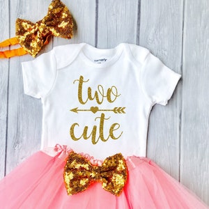 May include: A pink tulle tutu skirt with a gold glitter bow, a white onesie with the text "two cute" in gold glitter, and a gold glitter headband with a bow.
