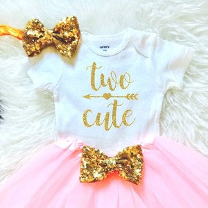 May include: A pink tulle tutu skirt with a gold glitter bow. A white onesie with gold glitter text that says "two cute" with a gold glitter arrow and heart. A gold glitter headband with a bow.