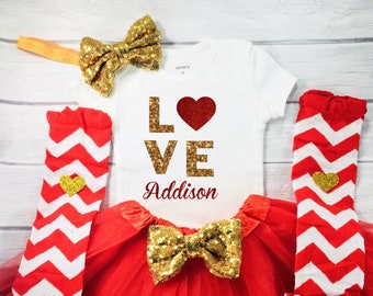 May include: A baby outfit set featuring a white onesie with the word "LOVE" in gold glitter and a red heart. The set includes a red tutu, gold sequin bow, red and white chevron leg warmers, and a gold headband.