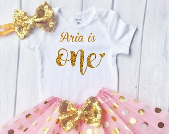 May include: A pink tulle tutu skirt with gold polka dots and a gold sequin bow. A white onesie with gold glitter lettering that says "Aria is one" and a gold sequin bow headband.