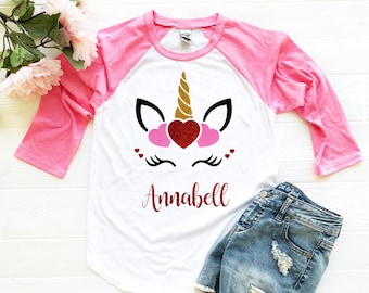 Könnte beinhalten: Ein weißes und pinkfarbenes Baseball-T-Shirt mit einem Unicorn-Gesichtsmotiv. Das Einhorn hat ein goldenes Horn, rosa Ohren und rote Glitzerherzen. Der Name "Annabell" ist in rotem Glitzer unter dem Einhorn geschrieben.