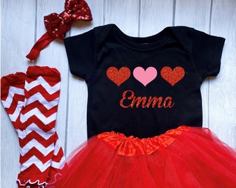 May include: A red and black baby outfit with a black bodysuit, red tutu skirt, and red and white leg warmers. The bodysuit has three glitter hearts, one pink and two red, and the name "Emma" written in red glitter.