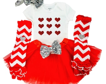May include: A red and white chevron leg warmer with a silver heart, a red tulle skirt with a silver sequin bow, a white bodysuit with red glitter hearts, and a silver bow headband.