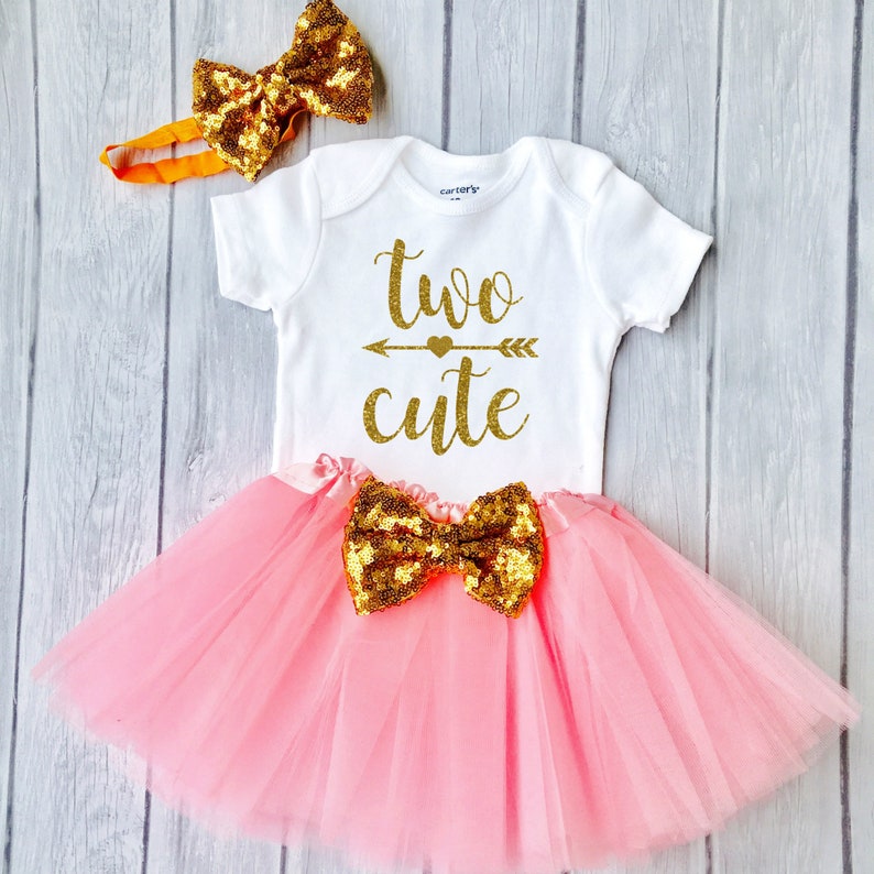 May include: A pink tulle tutu skirt with a gold glitter bow, a white onesie with the text "two cute" in gold glitter, and a gold glitter headband with a bow.