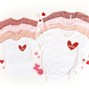 May include: Two white t-shirts with red glitter hearts. The larger shirt says "MAMA" and the smaller shirt says "MINI". The shirts are surrounded by pink and red hearts.