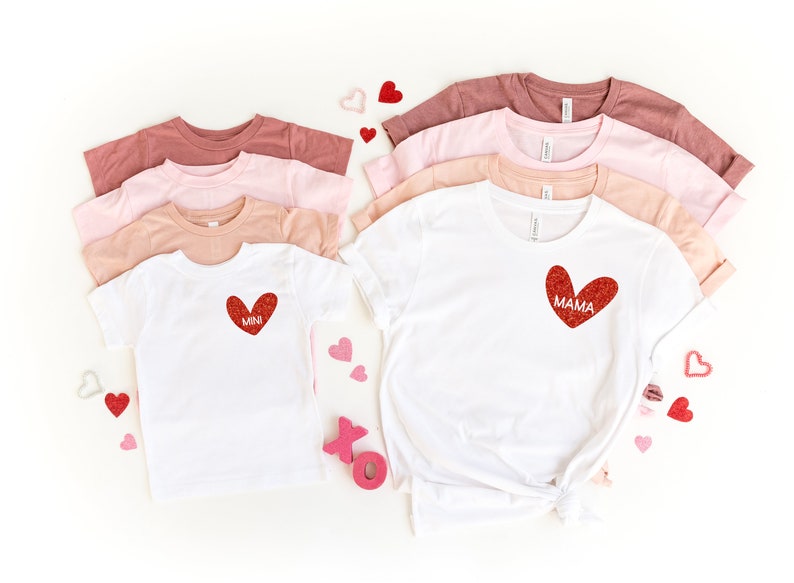 May include: Two white t-shirts with red glitter hearts. The larger shirt says "MAMA" and the smaller shirt says "MINI". The shirts are surrounded by pink and red hearts.