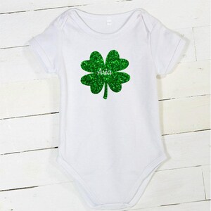 May include: White baby bodysuit with a green glitter shamrock and the name "Aria" printed on it.