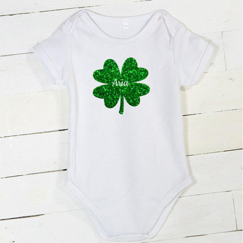 May include: White baby bodysuit with a green glitter shamrock and the name "Aria" printed on it.