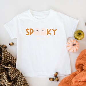 May include: White t-shirt with a brown "Spooky" graphic featuring three ghosts. The shirt is surrounded by fall-themed decor, including acorns and small pumpkins.