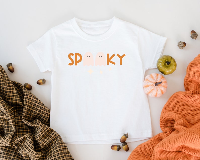 May include: White t-shirt with a brown "Spooky" graphic featuring three ghosts. The shirt is surrounded by fall-themed decor, including acorns and small pumpkins.