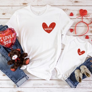 May include: Two white t-shirts with red glitter hearts. The larger shirt says "MAMA" and the smaller shirt says "MINI". The shirts are surrounded by red heart-shaped balloons, a teddy bear, and jeans.