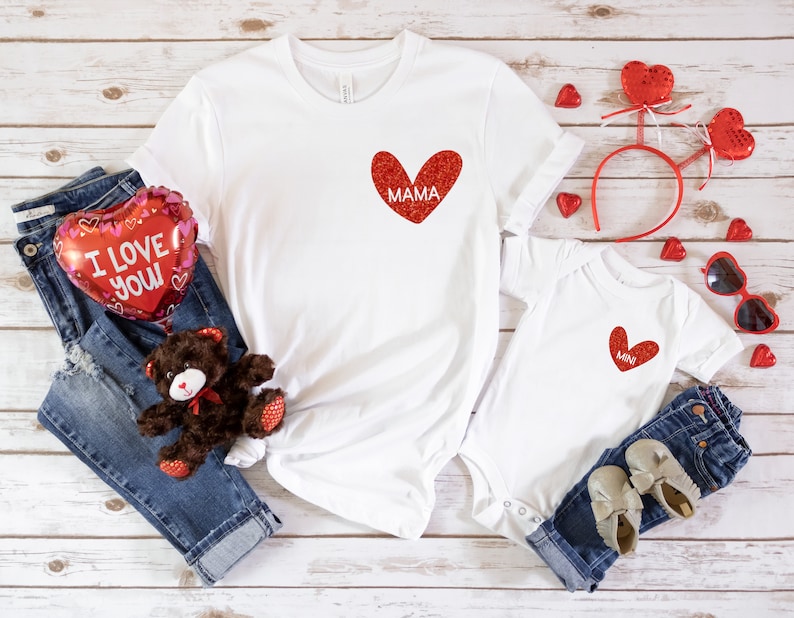 May include: Two white t-shirts with red glitter hearts. The larger shirt says "MAMA" and the smaller shirt says "MINI". The shirts are surrounded by red heart-shaped balloons, a teddy bear, and jeans.