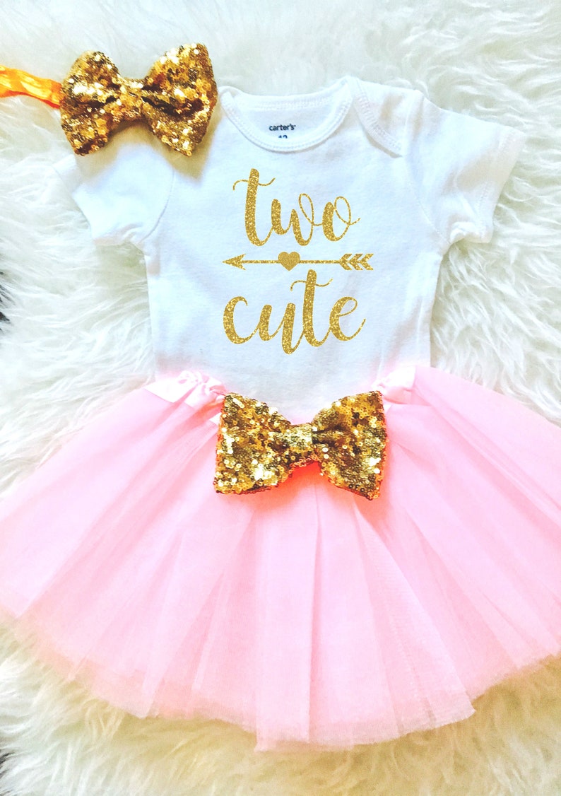 May include: A pink tulle tutu skirt with a gold glitter bow. A white onesie with gold glitter text that says "two cute" with a gold glitter arrow and heart. A gold glitter headband with a bow.