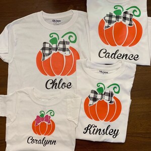 May include: Four white t-shirts with a pumpkin design and a name on each. The pumpkins have a black and white checkered bow. The names are Coralyann, Chloe, Kinsley, and Cadence.